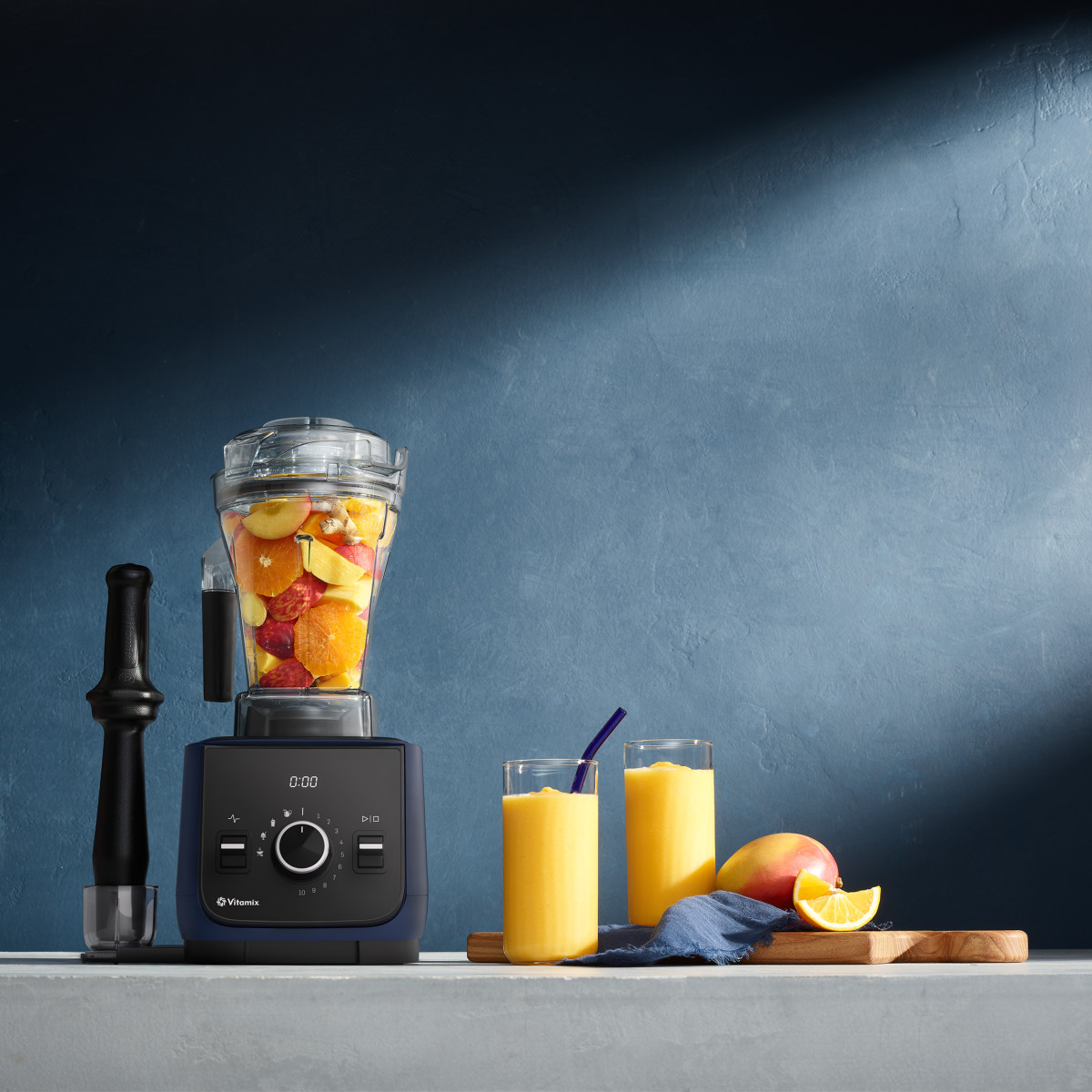 vitamix mixer with tamper with glasses of fruit smoothies staged next to it