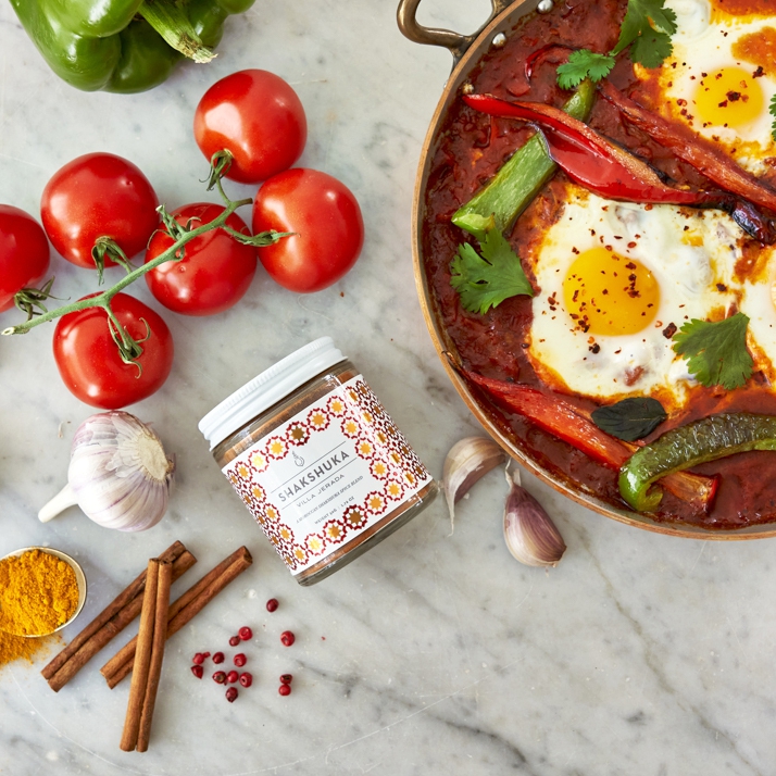Shaksuka jar next to fresh tomatoes, fried eggs and garlic